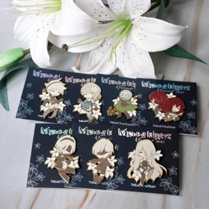 May include: Enamel pins featuring anime-style characters with floral accents. Each pin is a different character with varying color schemes, set against a dark background with white floral patterns. The text "THEARTTHATRAN" is visible on the cards.