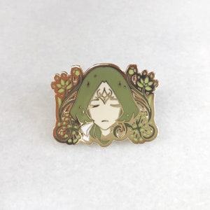 May include: A gold-coloured enamel pin depicting a calm face with closed eyes, green hair, and a small crown. The pin is framed by floral and leaf patterns in green and gold. The design has a fantasy or mythological theme.