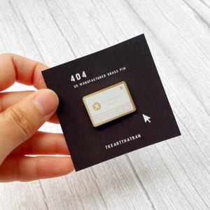Pode incluir: Um alfinete dourado com fundo branco que apresenta uma mensagem de erro de computador que diz "Catastrophic Failure" com um X vermelho. O alfinete está em um cartão preto com o texto "404 UK MANUFACTURED BRASS PIN" e "THE ARTTHATRAN".