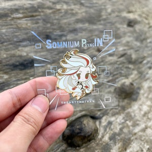 May include: A gold-colored enamel pin featuring a white-haired character with red accents. The character is wearing a white and red outfit and has a white halo. The pin is on a clear background with the text "Somnium Psyc. IN" and "TheArtThatTran" in gold.