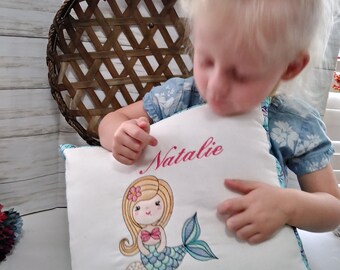 May include: White pillow with a mermaid design embroidered on it. The mermaid is light blue and green with pink hair and a pink flower. The pillow also has the name "Natalie" embroidered on it in pink.