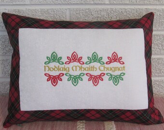 May include: Rectangular pillow with a red and black plaid pattern border. The center features a white panel with embroidered green and red Celtic knot designs and the text "Nollaig Mhaith Chugnat" in gold.