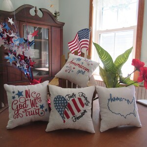 May include: Four decorative throw pillows with patriotic themes. The pillows are white with red, blue, and white embroidery. The pillows feature the phrases "In God We Trust", "Faith Family Freedom", "All American Girl", and "Home".