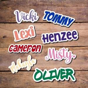 May include: A collection of name stickers in various colors and fonts, including "Vicki," "Tommy," "Lexi," "Henzee," "Cameron," "Misty," "Makayla," and "Oliver." Each sticker has a white border and is set against a wooden background.