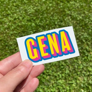 May include: A white sticker with the name "GENA" in large, colorful, 3D block letters. The letters are yellow with a pink outline and a blue shadow. The sticker is held against a background of green grass.
