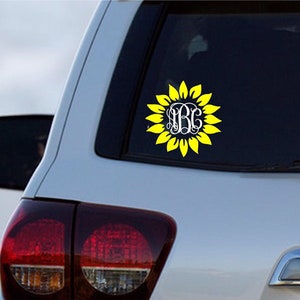 Puede incluir: Una calcoman&iacute;a de girasol amarilla con un monograma blanco en el centro. La calcoman&iacute;a est&aacute; en la ventana trasera de un coche plateado.