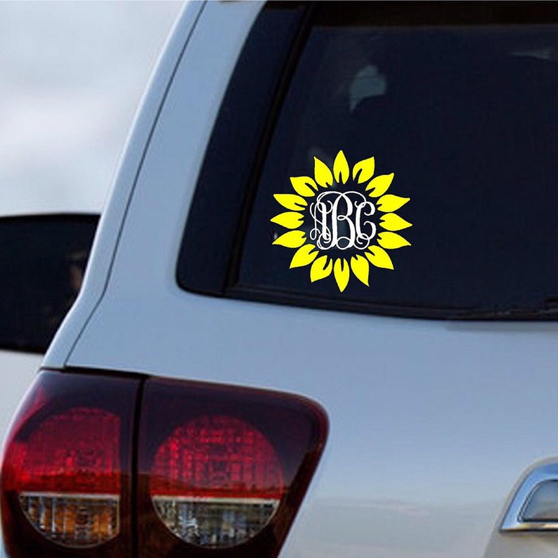 Puede incluir: Una calcoman&iacute;a de girasol amarilla con un monograma blanco en el centro. La calcoman&iacute;a est&aacute; en la ventana trasera de un coche plateado.