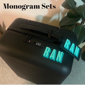 May include: Black suitcase with the text "Monogram Sets" at the top. The suitcase has a combination lock and a luggage tag that says "RAN" in turquoise letters. The suitcase is sitting on a light gray surface.