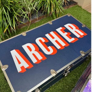 May include: A rectangular navy blue storage trunk featuring the name ARCHER printed in large white letters with a bold orange drop shadow. The trunk is constructed with silver-toned metal corner reinforcements and metal latches along the edges.