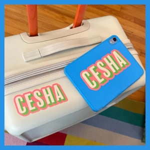 May include: A white suitcase with a pink, green, and white sticker that says "CESHA" on the front. A blue case with a pink, green, and white sticker that says "CESHA" is attached to the handle.