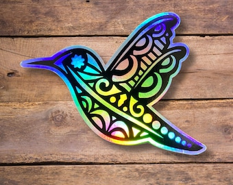May include: A holographic iridescent sticker of a hummingbird with a detailed, intricate design. The hummingbird is flying with its wings spread out. The sticker is on a wooden background.
