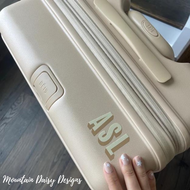 Op de afbeelding: Beige koffer met de letters "ASL" in lichtgroen. De koffer heeft een rits en een handvat. De merknaam "B&Eacute;IS" is zichtbaar op het handvat en de zijkant van de koffer. De woorden "Mountain Daisy Designs" staan onderaan.