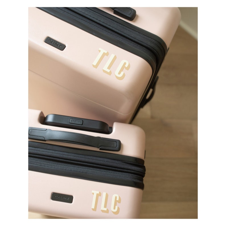 May include: Two blush pink CALPAK suitcases, one stacked on top of the other. Each suitcase has a black zipper and handle. The letters "TLC" are displayed on the front of each suitcase in a cream-colored font.