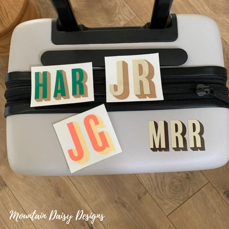May include: A light gray suitcase with a black zipper and handle. Four stickers with initials in various colors are placed on the suitcase. The stickers read HAR, JR, JG, and MRR. The text "Mountain Daisy Designs" is at the bottom.