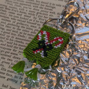 May include: A green, handmade keyring with a pixelated design featuring red, white, and black elements. The keyring has a silver clasp and green tassels. The word "Revolutionary" is visible in the bottom right corner.