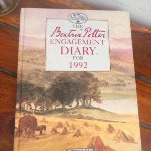 Può includere: Un libro con copertina rigida intitolato The Beatrix Potter Engagement Diary for 1992. La copertina presenta un'illustrazione ad acquerello di un paesaggio con covoni di fieno e alberi. Il testo include il logo Beatrix Potter Country World e F. Warne & Co.