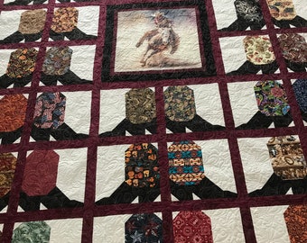May include: A quilt with a cowboy riding a bucking horse in the center. The quilt is made of various fabrics with different patterns and colors. The quilt is framed with a burgundy border.