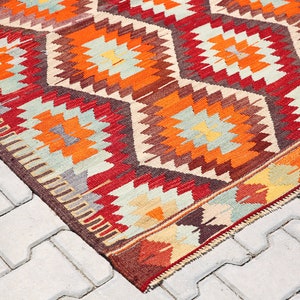 May include: A colorful, geometric patterned rug with red, orange, brown, and white colors. The rug is made of wool and has a traditional design.