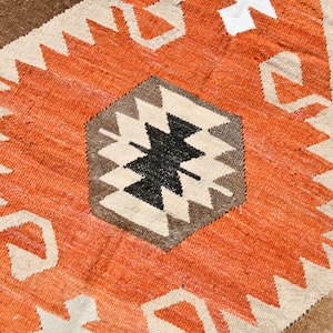 May include: A close-up of a woven rug with a geometric pattern in shades of brown, orange, white, and black. The rug has a textured surface and a traditional design.