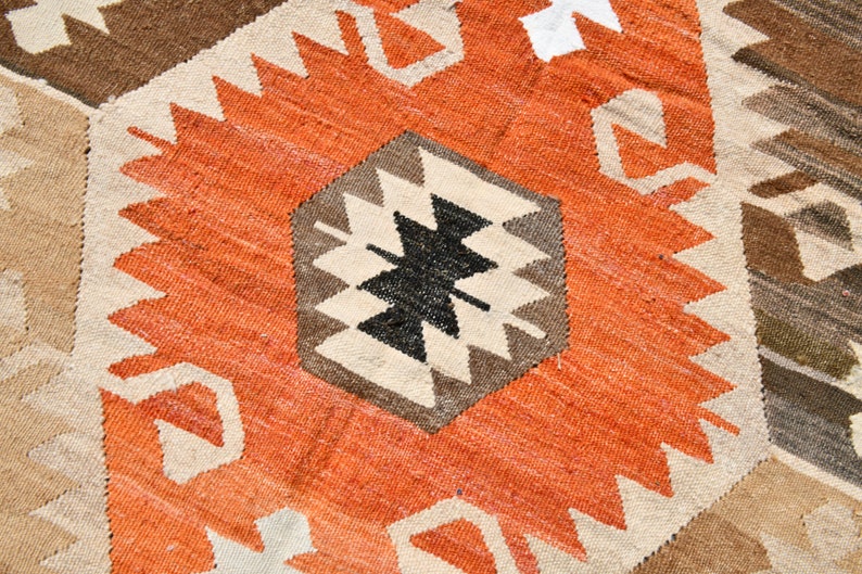 May include: A close-up of a woven rug with a geometric pattern in shades of brown, orange, white, and black. The rug has a textured surface and a traditional design.