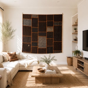 May include: A large, rectangular wall hanging featuring a patchwork design of dark brown, charcoal, and black fabric panels separated by thin, vibrant orange stitched lines, displayed prominently on a plain white wall above a white sectional sofa.