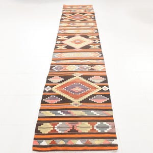 Puede incluir: Una alfombra larga y rectangular con un patrón geométrico. La alfombra presenta un diseño repetitivo de diamantes, rayas y otras formas en tonos de naranja, marrón, beige y morado. La alfombra probablemente está hecha de tela tejida.
