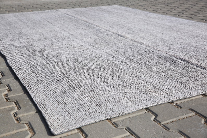 May include: A gray and white woven rug with a textured surface. The rug is laid out on a gray concrete surface.
