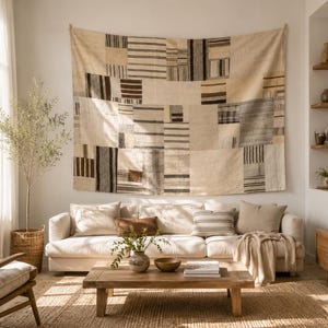 May include: A large rectangular wall hanging featuring a patchwork design of cream, beige, brown, and charcoal gray fabric panels with various striped and solid patterns, displayed horizontally above a white sofa in a living room setting.
