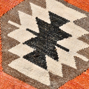 May include: Close-up of a woven rug with a geometric pattern in shades of brown, black, and white. The pattern features a central star shape with pointed edges. The rug is surrounded by a border of orange fabric.