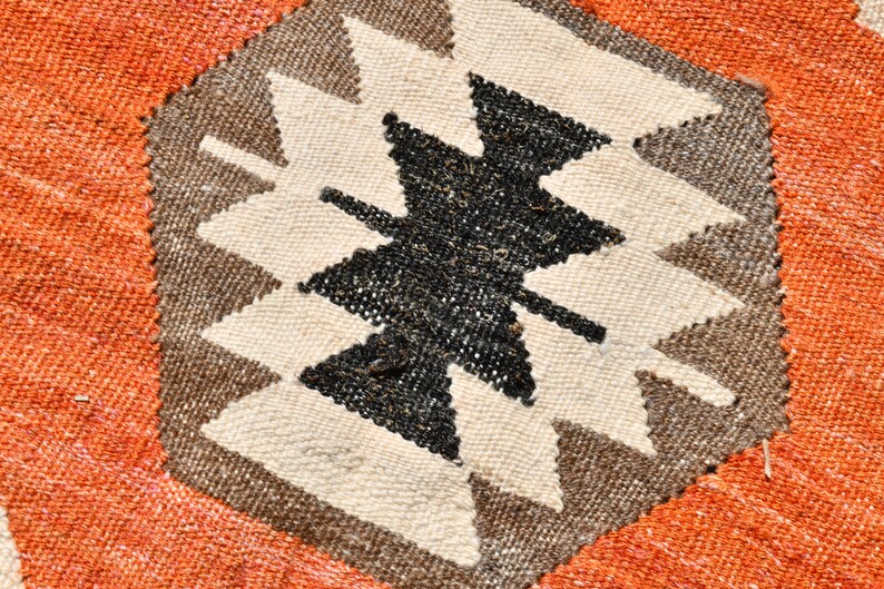 May include: Close-up of a woven rug with a geometric pattern in shades of brown, black, and white. The pattern features a central star shape with pointed edges. The rug is surrounded by a border of orange fabric.