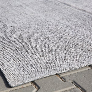 May include: A close-up of a gray and white woven rug with a textured surface. The rug is lying on a gray concrete surface.