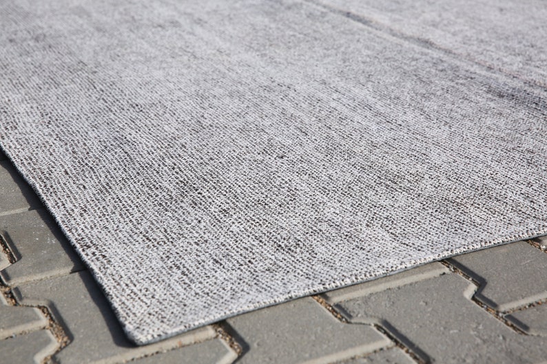 May include: A close-up of a gray and white woven rug with a textured surface. The rug is lying on a gray concrete surface.