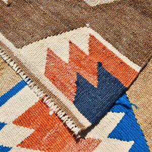 May include: Close-up of a woven rug with a geometric pattern in shades of brown, white, blue, and orange. The rug has a fringe border.