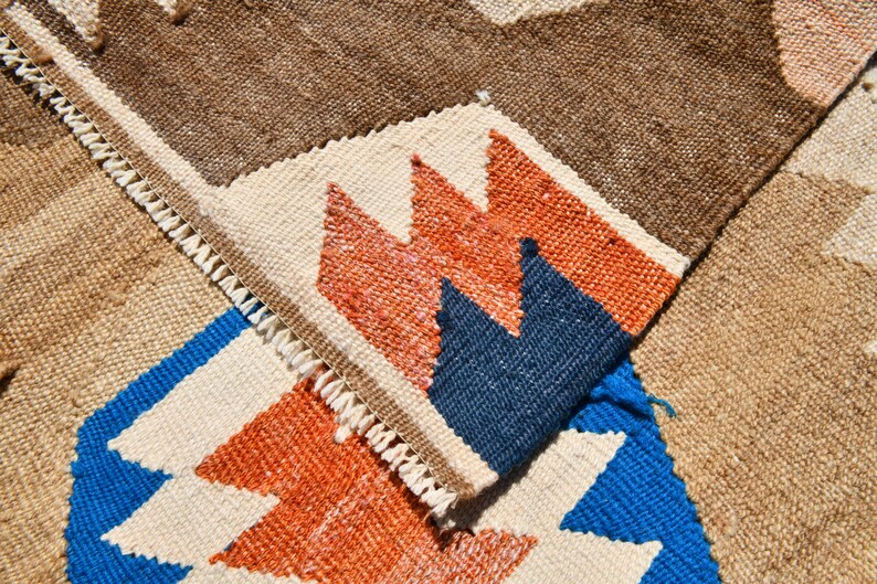 May include: Close-up of a woven rug with a geometric pattern in shades of brown, white, blue, and orange. The rug has a fringe border.