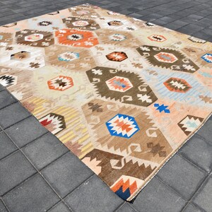 May include: A vintage Turkish rug with a geometric pattern in shades of brown, orange, blue, and white. The rug has a distressed look and is laid out on a gray tiled floor.