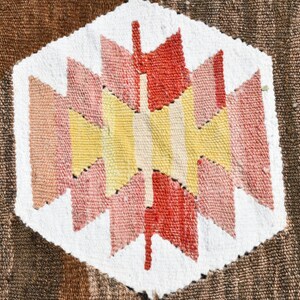 May include: A close-up of a woven rug with a geometric pattern in shades of red, pink, yellow, and white. The pattern is centered on a white background and surrounded by a brown background.
