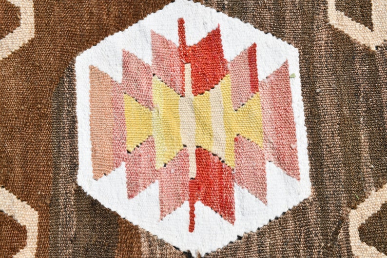 May include: A close-up of a woven rug with a geometric pattern in shades of red, pink, yellow, and white. The pattern is centered on a white background and surrounded by a brown background.