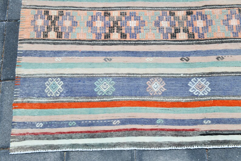 May include: A handwoven rug with a striped pattern in shades of blue, orange, green, and white. The rug has a geometric design with small squares and diamonds.