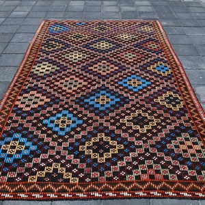 Può includere: Un tappeto turco vintage con un motivo geometrico in tonalità di rosso, blu, giallo e nero. Il tappeto ha un bordo con frange ed è tessuto con dettagli intricati.