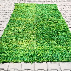 May include: A rectangular, shaggy rug with a vibrant green color. The rug has a thick, textured pile, and the color varies from light to dark green. The rug is placed on a gray, patterned surface.