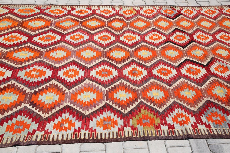 May include: A traditional Turkish rug with a geometric pattern in shades of red, orange, beige, and brown. The rug has a fringe border.