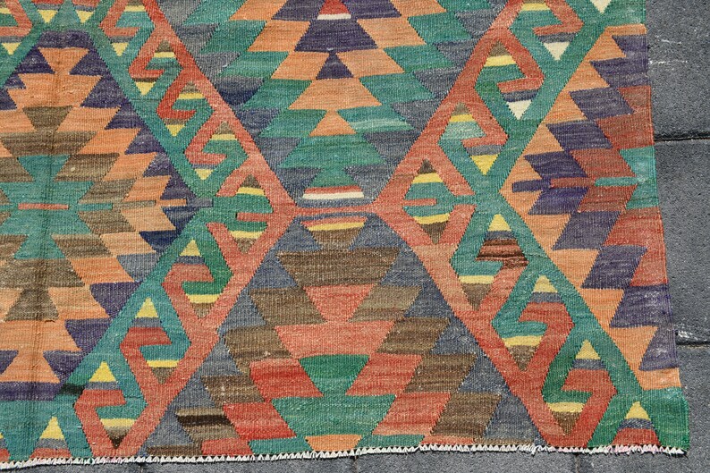 Pu&ograve; includere: Un tappeto colorato con un motivo geometrico di triangoli in tonalit&agrave; di blu, verde, arancione e marrone. Il tappeto ha un bordo con frange.