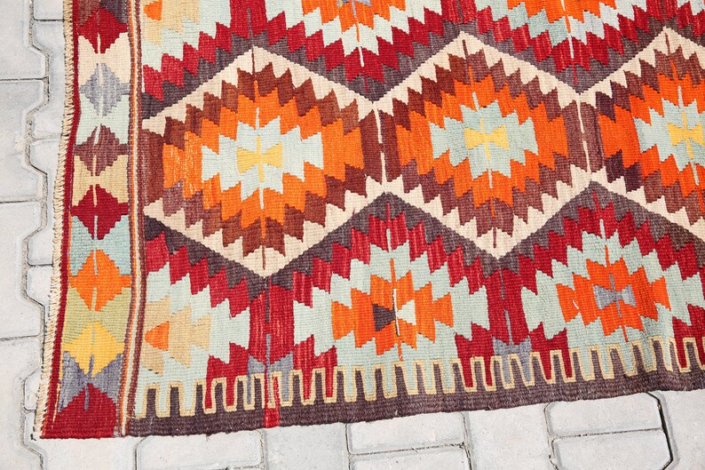 May include: A colorful rug with a geometric pattern in shades of red, orange, brown, and beige. The rug has a fringe border.