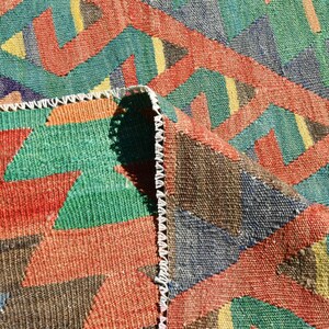 Pu&ograve; includere: Primo piano di un tappeto tessuto colorato con un motivo geometrico in tonalit&agrave; di rosso, verde, blu e marrone. Il tappeto ha un bordo con frange.