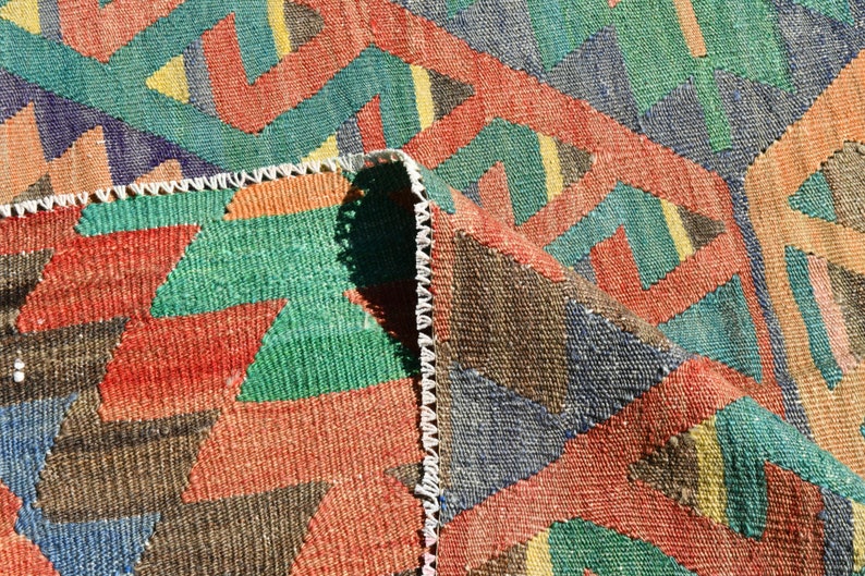 Pu&ograve; includere: Primo piano di un tappeto tessuto colorato con un motivo geometrico in tonalit&agrave; di rosso, verde, blu e marrone. Il tappeto ha un bordo con frange.