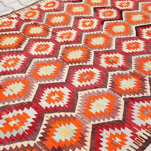 May include: A traditional Turkish kilim rug with a geometric pattern in shades of red, orange, beige, and brown. The rug features a repeating diamond shape with a starburst design in the center.