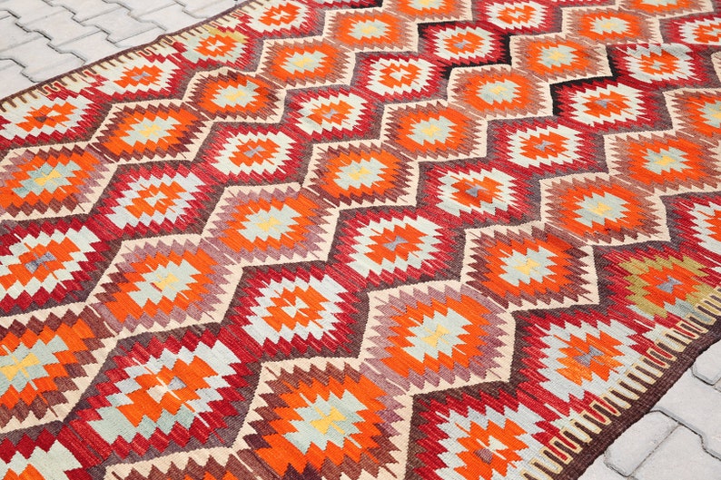 May include: A traditional Turkish kilim rug with a geometric pattern in shades of red, orange, beige, and brown. The rug features a repeating diamond shape with a starburst design in the center.