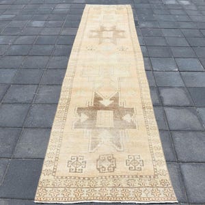 Könnte beinhalten: Ein rechteckiger, beigefarbener Läufer mit einem zentralen geometrischen Muster in Brauntönen. Der Teppich hat ein sich wiederholendes Bordürendesign und liegt auf einer dunkelgrauen Fliesenoberfläche. Das Design des Teppichs ist symmetrisch und traditionell.