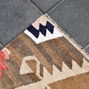 May include: A close-up of a woven rug with a geometric pattern in shades of brown, beige, white, red, and blue. The rug has a fringe border.