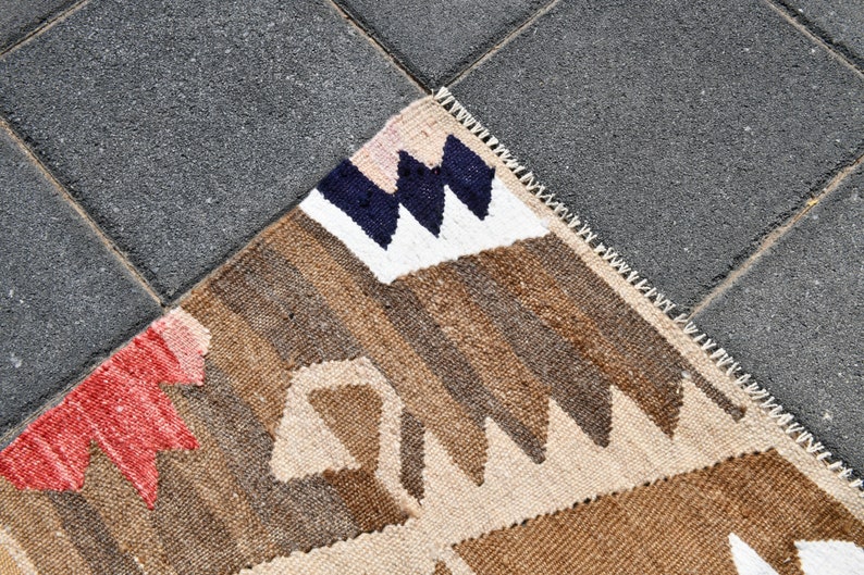 May include: A close-up of a woven rug with a geometric pattern in shades of brown, beige, white, red, and blue. The rug has a fringe border.